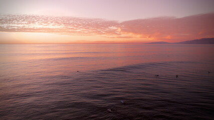 Aerial view of the calm ocean waters reflecting a vibrant sunset with surfers enjoying the waves, San Francisco, California, United States.