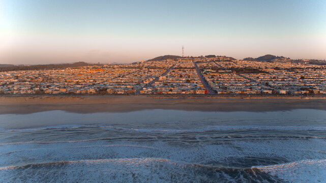 Aerial view of a vibrant sunset casting long shadows across the Sunset District's neat rows of houses, meeting the foamy waves of the Pacific, San Francisco, California, United States.