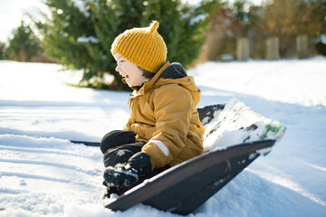 Laughing child in mustard jacket and yellow hat rides in a snow shovel, sitting low in the snow. Improvised winter sledding, playful spontaneity in cold sunlight.