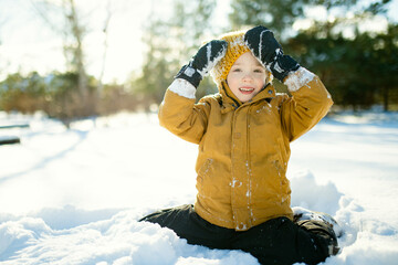 Smiling child in mustard jacket and knit hat crouches in snow, cheeks rosy from the cold. Winter...