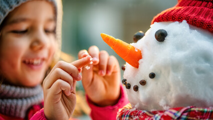 Child joyfully decorating a snowman with a carrot nose, pebble eyes and a red knit hat, adding tiny snow details in warm winter light with a soft festive bokeh background