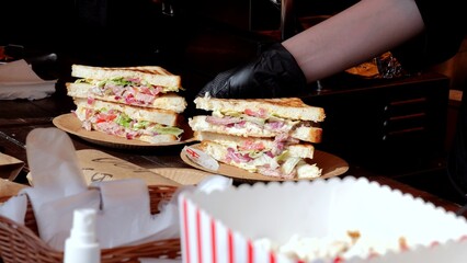 Vendor crafts multitiered deli sandwiches, Food stand displays stacked sandwiches with condiments and popcorn, Bustling street market scene with vendor preparing multilayer sandwiches and snack boxes