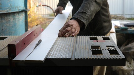 A worker in dark clothes operates a circular saw and cuts a long thin plank into two pieces. A carpenter works in a workshop at a sawing machine during the processing of wood material.