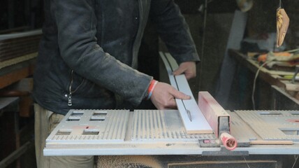 An impersonal carpenter works on a circular saw, cutting a narrow wooden plank lengthwise against the background of an equipped workshop, machine processing and preparation of panels for construction 