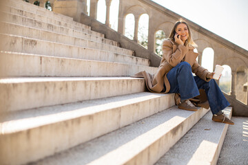 Woman enjoying coffee and talking on phone while sitting on steps in sunny outdoor setting