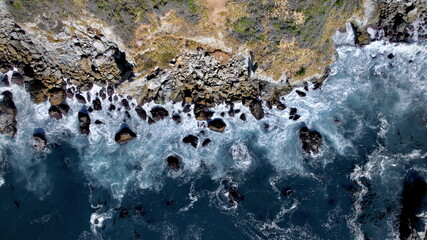 Aerial view of waves crashing against the rocky cliffs of the Big Sur coastline, contrasting the deep blue sea with the rugged terrain, Big Sur, California, United States.