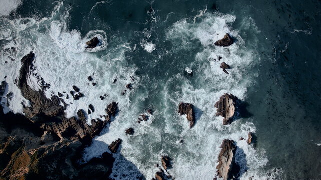Aerial view of the rugged coastline where the dark, jagged rocks meet the turquoise sea and foamy white waves crash dramatically against the shore, Big Sur, California, United States.