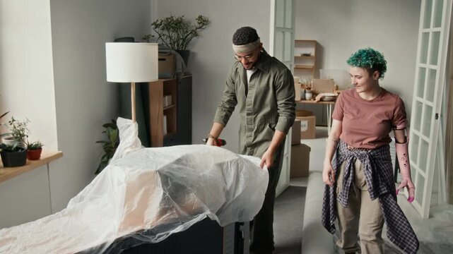 Young man covering furniture with protective film, girlfriend with bionic arm walking up to him while preparing for redecorating apartment