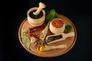 Round wooden tray with assorted spices including turmeric, cardamom, chili, pepper, garlic, mint on black background