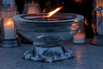 Duża, dekoracyjna świeca płonąca na nagrobku w Dzień Wszystkich Świętych (Zbawionych) wieczorem. Główna świeca umieszczona jest w ozdobnym szklanym świeczniku o szerokiej, płytkiej,misternej  misie  © Grzegorz