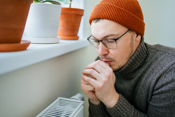Man wearing warm hat and sweater suffering from cold at home, paying expensive heating bills and saving money during energy crisis, warming hands near radiator due to high costs
