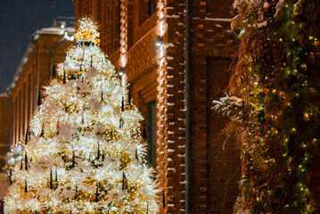 Brightly lit christmas tree sparkling with warm white lights and draped in snow stands outside a brick building, creating a festive winter scene during a snowing holiday evening