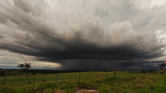 A storm over a farm field, seen in the distance, approaches rapidly with extremely heavy clouds on a late afternoon in Brazil.