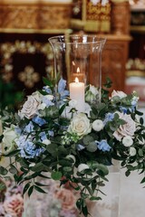 Floral centerpiece featuring blush and white roses, blue delphiniums, and green foliage with a burning candle in a glass vase, used for wedding decor