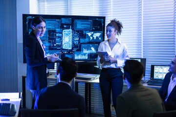 Female executive leads a crucial discussion, presenting complex data and cybersecurity analysis on screen to diverse team of analysts at command center. Global business strategy, financial technology