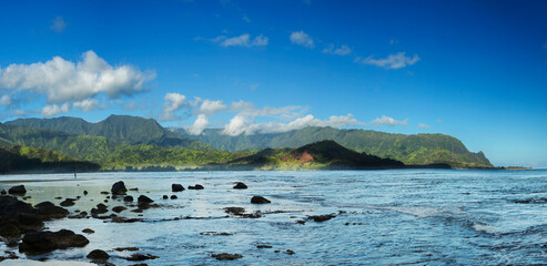 A panorama of Hanalei Bay on the north shore of Kauai, Hawaii., with black volcanic boulders in the foreground.