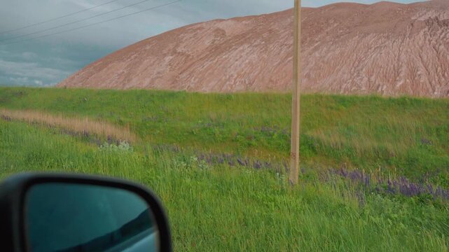 Personal perspective driving along a quiet rural road, passing a large industrial slag heap from a mining operation under a bright sky, illustrating travel through a modified landscape