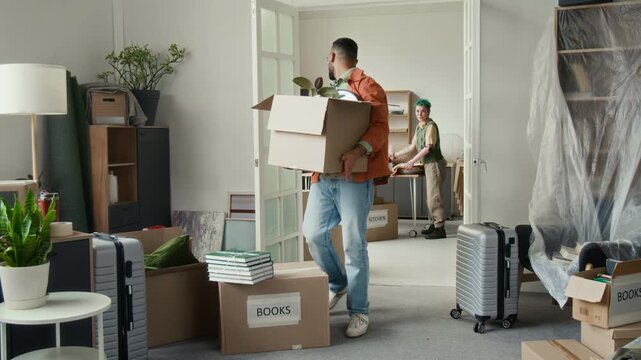 Young man carrying cardboard box and looking back speaking to his girlfriend, then he stumbling over another box and hitting his leg during moving process