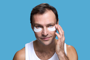 Portrait of Caucasian middle aged man applying under eye patches, looking into camera, gently touching face with hand, demonstrating skincare routine against blue background