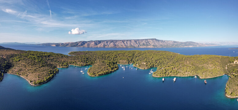 Aerial view of azure waters embrace the verdant, tree-covered coastlines, where boats rest peacefully, with distant mountains under a vast blue sky, Stari Grad, Split-Dalmatia County, Croatia.