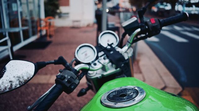 A shallow focus shot of a green motorcycle's handlebars and controls, with raindrops on the mirror and tank