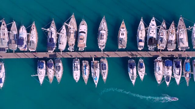 Top down aerial view captures a large marina green water many sailboats docked in perfect, orderly rows. Drone overhead shot of sailboats neatly docked along symmetrical wooden pier Greece