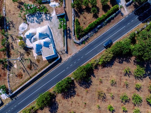 Aerial view of a solitary car glides along a sun-drenched road dividing rustic fields and traditional whitewashed dwellings, Monopoli, Apulia, Italy.