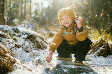 Child in yellow hat crouches on frozen forest marsh, reaching toward grass trapped under clear ice. Intimate winter exploration, early childhood wonder in northern nature. © MNStudio