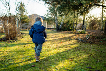 Obraz premium Smiling child in hoodie and puffer jacket stands in a green garden on a grey day. Mild European winter or late autumn, quiet moment between seasons.