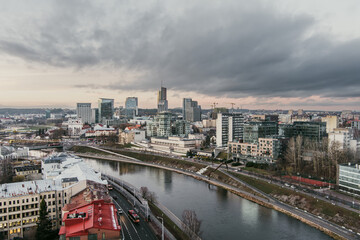 Aerial view of Vilnius cityscape on a winter day, with modern high-rises along the Neris River under cloud cover. Urban contrast of glass architecture, historic rooftops, and season stillness.