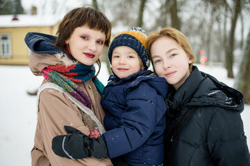 Two teenage girls embrace their younger brother in a snowy park, dressed in winter coats. A tender family moment in cold weather, warmth held close despite the chill.