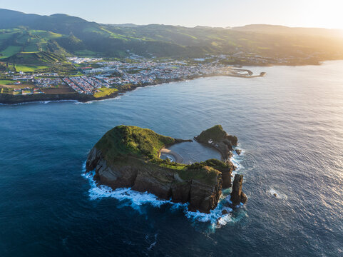 Aerial view of the rugged islet of Vila Franca do Campo emerges from the deep blue Atlantic, juxtaposed against the town and verdant hills, Vila Franca do Campo, Azores, Portugal.