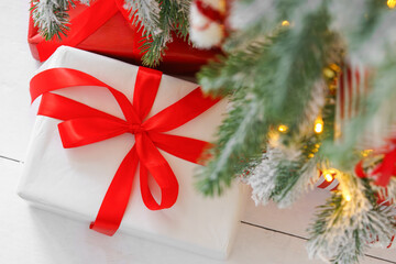 Beautiful present, gift box wrapped in elegant white paper with red bow under green snowy festive Christmas tree with warm lights. Cozy winter holiday atmosphere perfect for seasonal celebrations