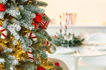Frosted Christmas tree decorated with sparkling lights and festive red ornaments on blurred dining background with holiday setting. Festive atmosphere of winter holidays and celebrations.