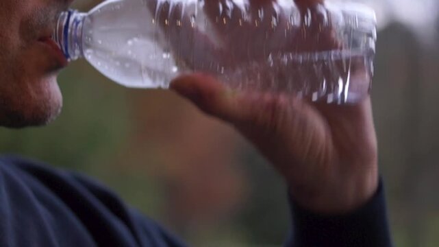 Man hydrating with water from a disposable, single-serving plastic bottle against a natural, blurred background. Represents the global reliance on plastic packaging