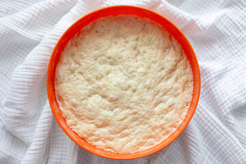 Risen yeast dough in an orange bowl on a white textured cloth. Soft, airy surface showing fermentation bubbles, ideal for baking, homemade bread, and culinary preparation themes.
