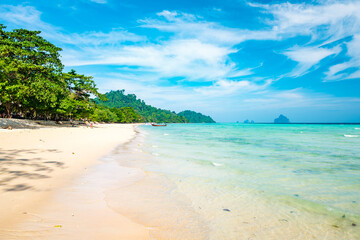 White sand tropical beach meeting clear turquoise water and lush green jungle, featuring a traditional longtail boat anchored offshore with distant islands under a bright blue sky © arkanto