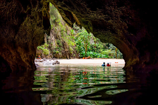 Swimmers exiting a dark sea cave, revealing a hidden light filled lagoon with a sandy beach and lush tropical vegetation on Koh Mook, Kantang, Trang, Thailand