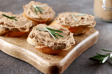 Chicken liver pate on toasted baguette slices garnished with rosemary on a wooden board, grey background
