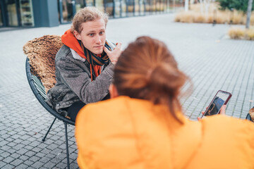 Mother with adult son sitting on an open terrace in a coffee shop, drinking coffee and having a lovely conversation. Mom shows interesting content on her smartphone in an over-the-shoulder portrait