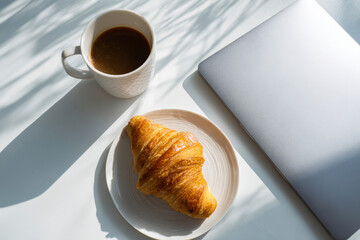 Morning ritual of sipping coffee and enjoying a fresh croissant near a laptop while sunlight fills the room with warmth and comfort