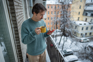 Thoughtful man standing on snowy balcony with hot coffee, checking smartphone. Pensive male scrolling phone outdoors on apartment balcony in winter morning, holding mug breathing frosty air. Slow life