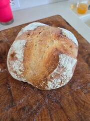 Freshly baked round loaf of bread with golden crust and flour dusting, placed on wooden cutting board with visible knife marks, cozy kitchen background.