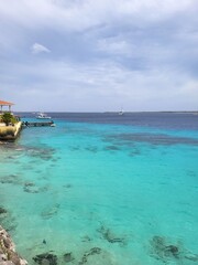 Obraz premium Turquoise water with anchored boats and sailboat, dock extending from land with yellow building and red roof, palm trees and clear sky, peaceful tropical coastal scene in natural light.