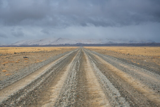 Gravel road stretching from Altai towards the Mongolian border with snowy mountain ridge. Desert steppe landscape under stormy cloudy sky showcasing wild remote terrain