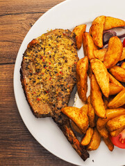 Fine dinner with spicy pork chop, potato wedges and fresh tomato on a plate on a wooden table. Tasty meal with meat and vegetables.