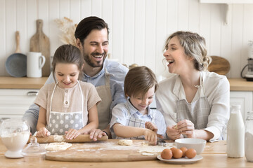 Baking with love. Joyful family of 4 wear aprons laugh bond making sweet cookies in bright cozy kitchen. Dad roll dough together with older daughter using pin as mom helps younger son cut fun shapes