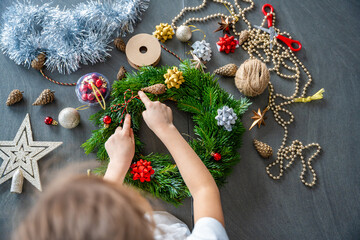 Little girl decorating a Christmas wreath while sitting at the table, top view. Capturing a...