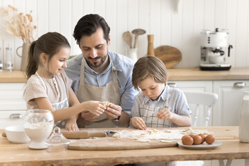 Daddy is best chef. Caring mid aged dad spend quality time with two kids in kitchen teach son daughter to knead roll dough to prepare tasty handmade cookies from natural ingredients using easy recipe