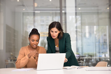 Two multiethnic female coworkers working together at laptop, pointing at screen, discussing task....